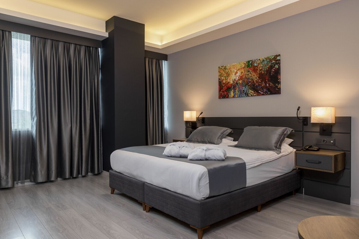 Quarto moderno com cama grande, cortinas cinzentas, pintura abstrata colorida e abajures.