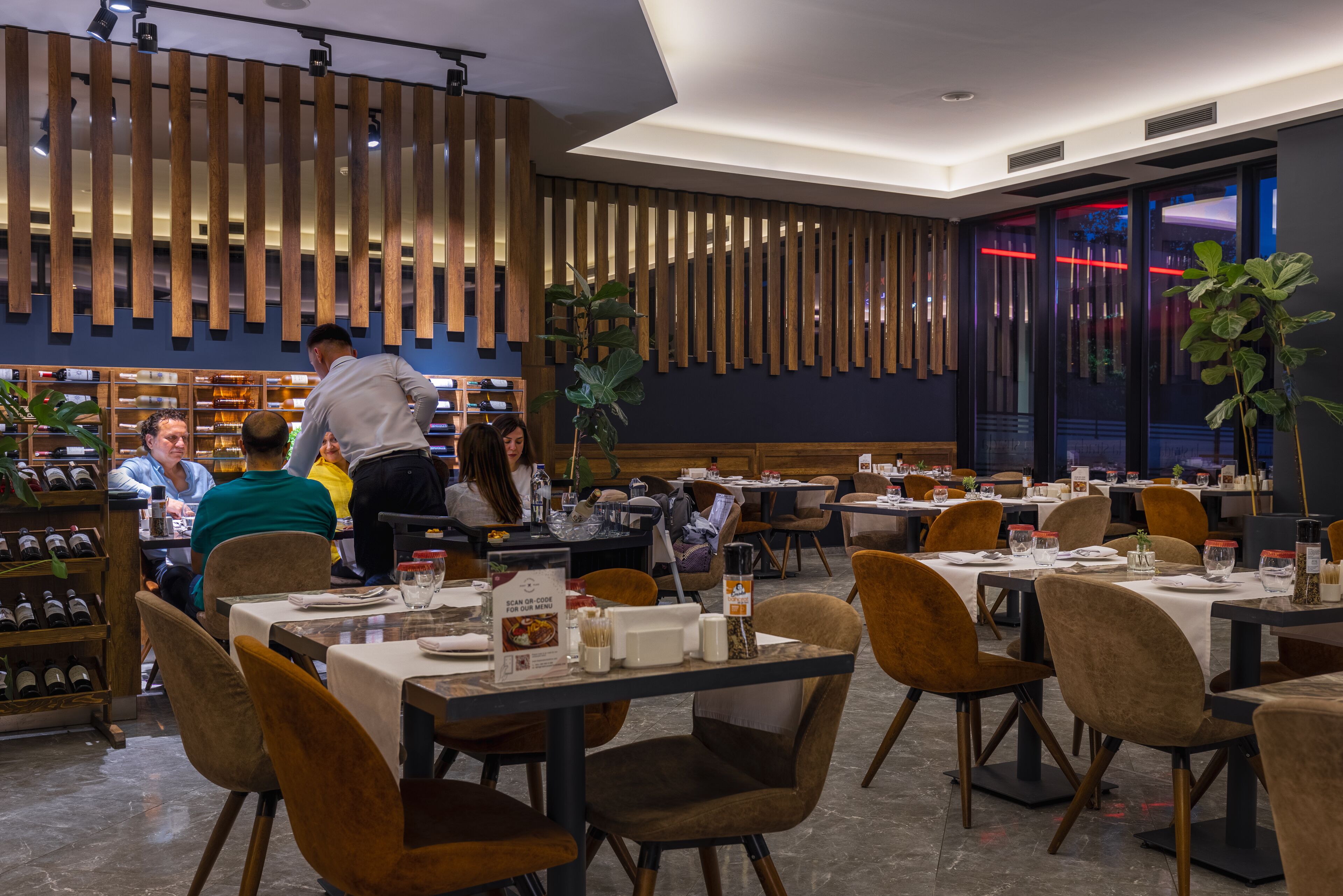 A modern restaurant interior with wooden slats on the walls, tables set with white tablecloths, and a variety of chairs.