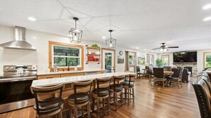 A modern kitchen with a central island, bar stools, and a dining area with a TV and fireplace. - Emory House 8 Bedroom Cabin by RedAwning (Pigeon Forge)