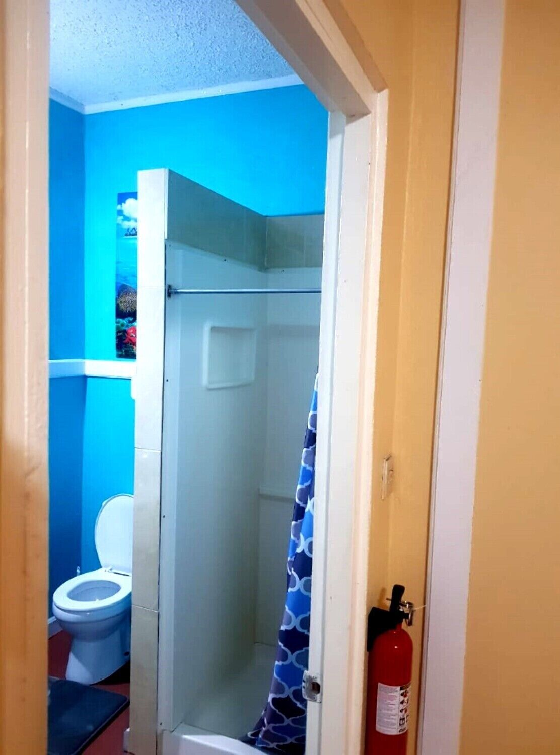 Ein Badezimmer mit blauer Wand, einer weißen Toilette, einer Dusche mit blau gemustertem Vorhang und einem an der Wand befestigten Feuerlöscher.