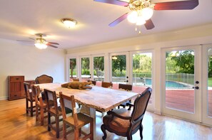 Un comedor espacioso con una mesa y sillas de madera, un ventilador de techo y ventanas amplias que dan a una piscina.