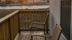 Een houten terras met twee stoelen, met uitzicht op een strand met huizen en palmbomen in de achtergrond tijdens de zonsondergang.