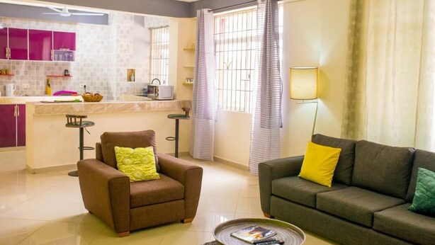 Sala de estar con sofá, sillón y mesa de centro. Hay una zona de cocina con armarios y una ventana con cortinas.