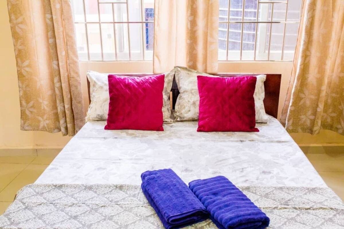 Un lit avec une couvre-lit à motifs, deux oreillers rouges et deux serviettes pliées posées sur une tête de lit blanche.