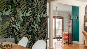 Un comedor con mesa de madera, sillas y papel tapiz estampado con helechos.