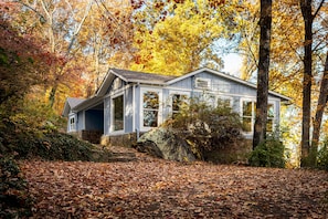 Un rustico chalet con fondamenta in pietra, ampie finestre e un tetto spiovente, immerso in un paesaggio autunnale con foglie sparse sul terreno.