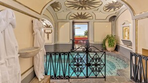 Un área de piscina interior lujosa con diseños ornamentales en el techo, una pileta de madera y una planta en maceta.