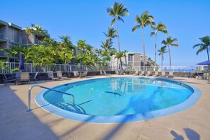 A swimming pool with lounge chairs and palm trees, surrounded by a fence and buildings.