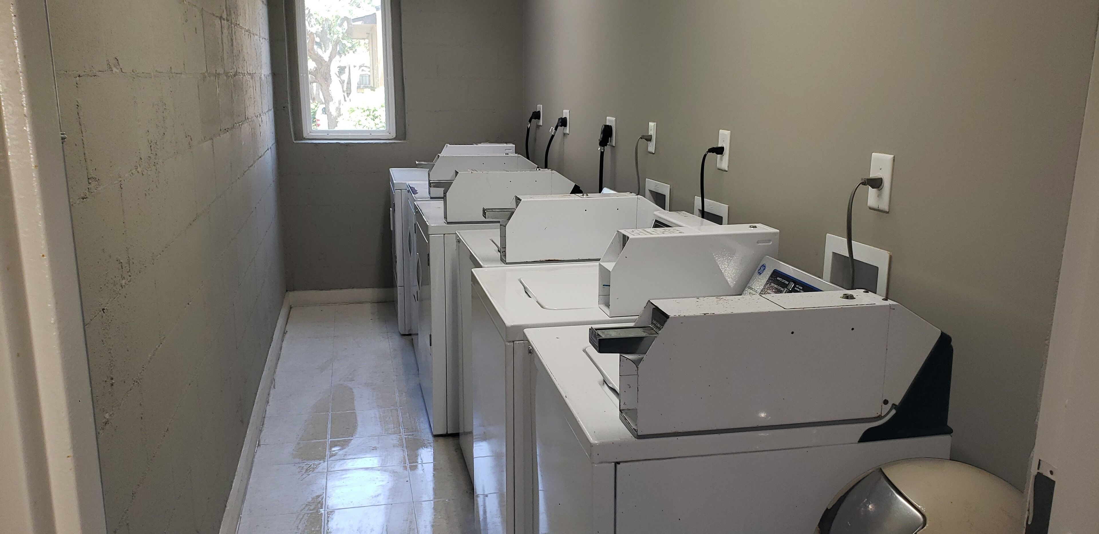 A laundry room with multiple washing machines lined up against the wall, a tiled floor, and a window providing natural light.
