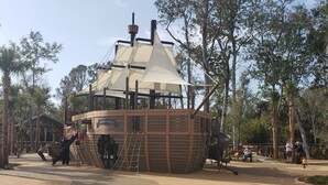 Una estructura de barco pirata de madera con una bandera, rodeada de árboles y suelo arenoso.