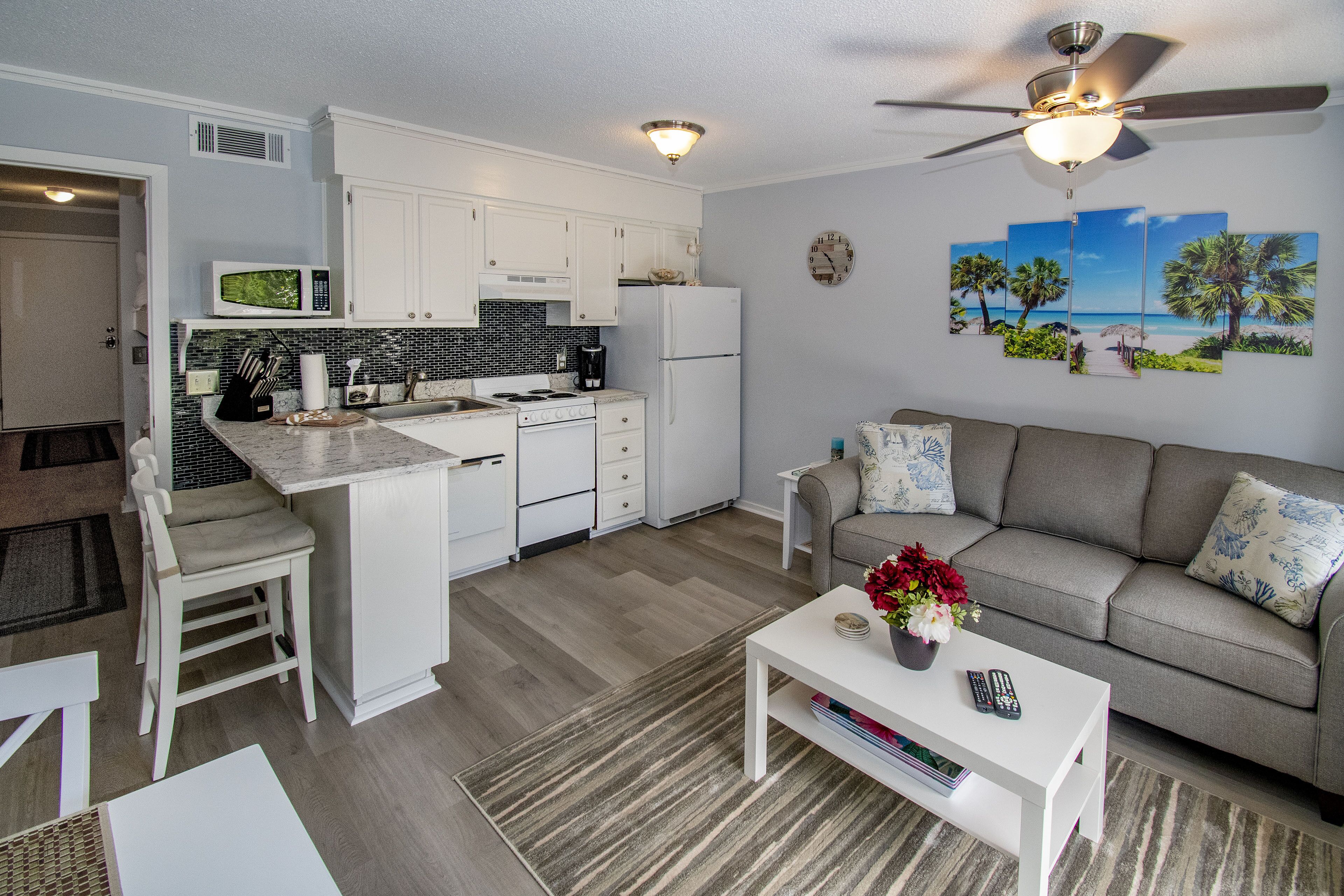 A modern kitchen with white cabinets, a microwave, and a refrigerator. There is a dining area with a white table and chairs. A gray sofa with decorative pillows is in the living room.