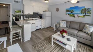 Una cocina moderna con armarios blancos, un microondas y un refrigerador. Hay un comedor con una mesa y sillas blancas. Un sofá gris con almohadones decorativos se encuentra en la sala.
