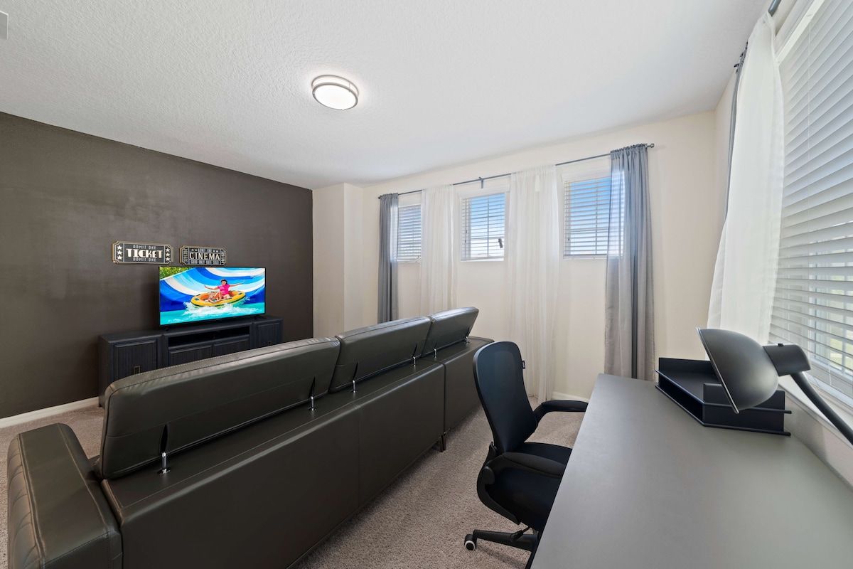 A modern living room with a flat-screen TV, a sofa, a desk with a chair, and a window with blinds.