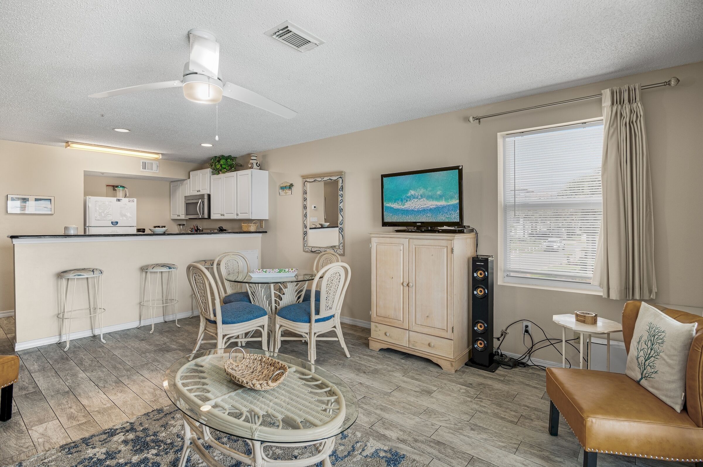 A compact living space with a kitchenette, a dining area with wicker furniture, a TV mounted on the wall, and a large window with curtains.