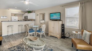 A compact living space with a kitchenette, a dining area with wicker furniture, a TV mounted on the wall, and a large window with curtains. - Pet-Friendly Gulf Front Condo • Free Beach Setup • 2 Pools + Hot Tub (Miramar Beach)