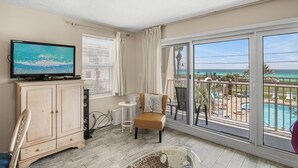 A balcony with a pool and ocean view, a television on a cabinet, and a chair with a cushion. - Pet-Friendly Gulf Front Condo • Free Beach Setup • 2 Pools + Hot Tub (Miramar Beach)
