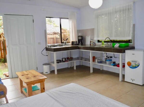 Une petite cuisine équipée d’un évier, d’une cuisinière et d’un réfrigérateur. On y trouve une fenêtre avec des rideaux, une table en bois et une chaise.