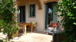 Une terrasse extérieure avec une table en bois et des chaises en osier, une petite table ronde avec une bougie, et une vue sur l’intérieur du bâtiment à travers une porte vitrée.