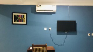 Ein Zimmer mit blauer Wand, einer Wand-Klimaanlage, einem gerahmten Bild, einem Fernseher und einem Holzstuhl mit kariertem Sitzkissen.