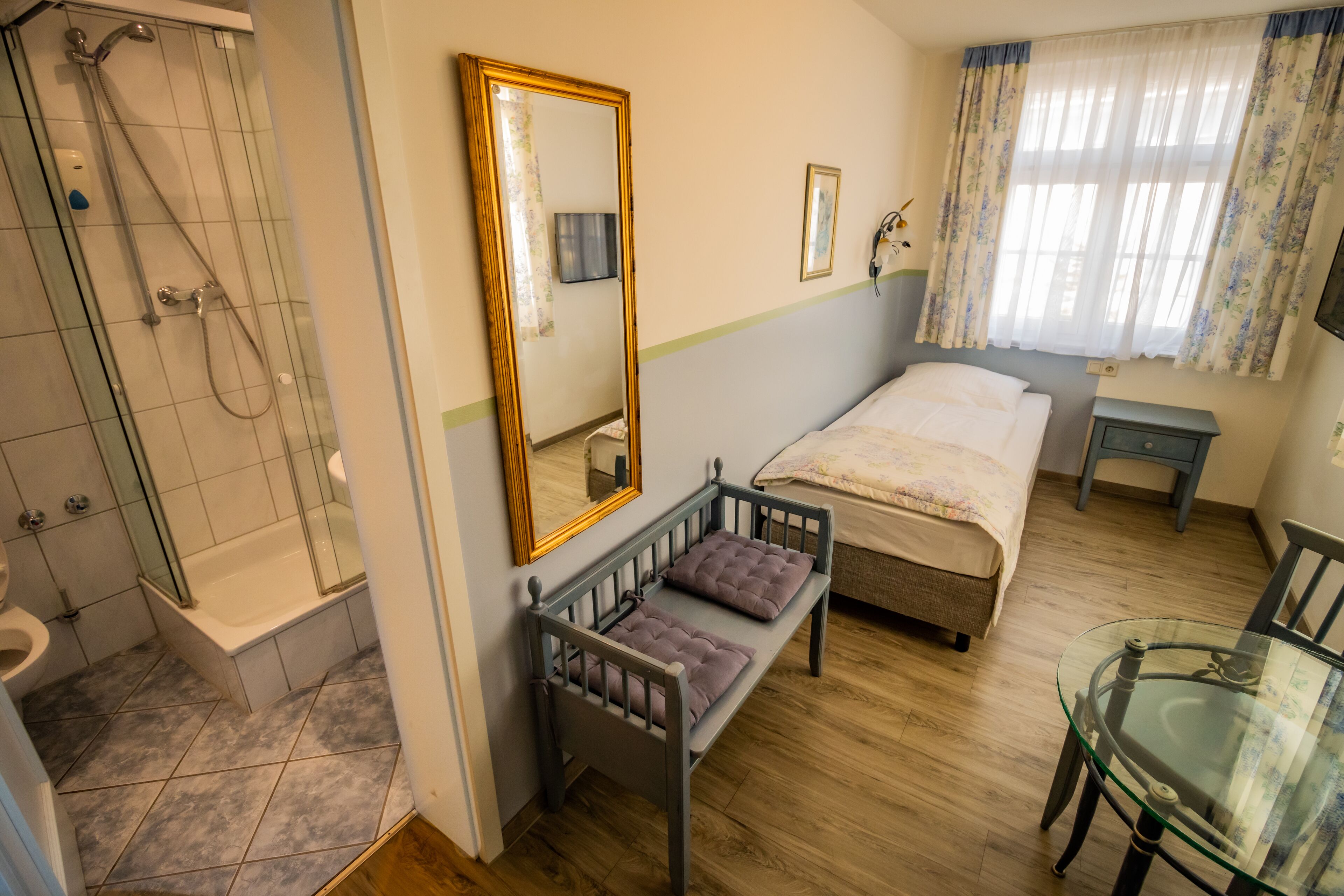 A bathroom with a shower, toilet, and sink. There is a bed with a pillow and a bedside table. A mirror hangs on the wall, and a window with curtains is visible.