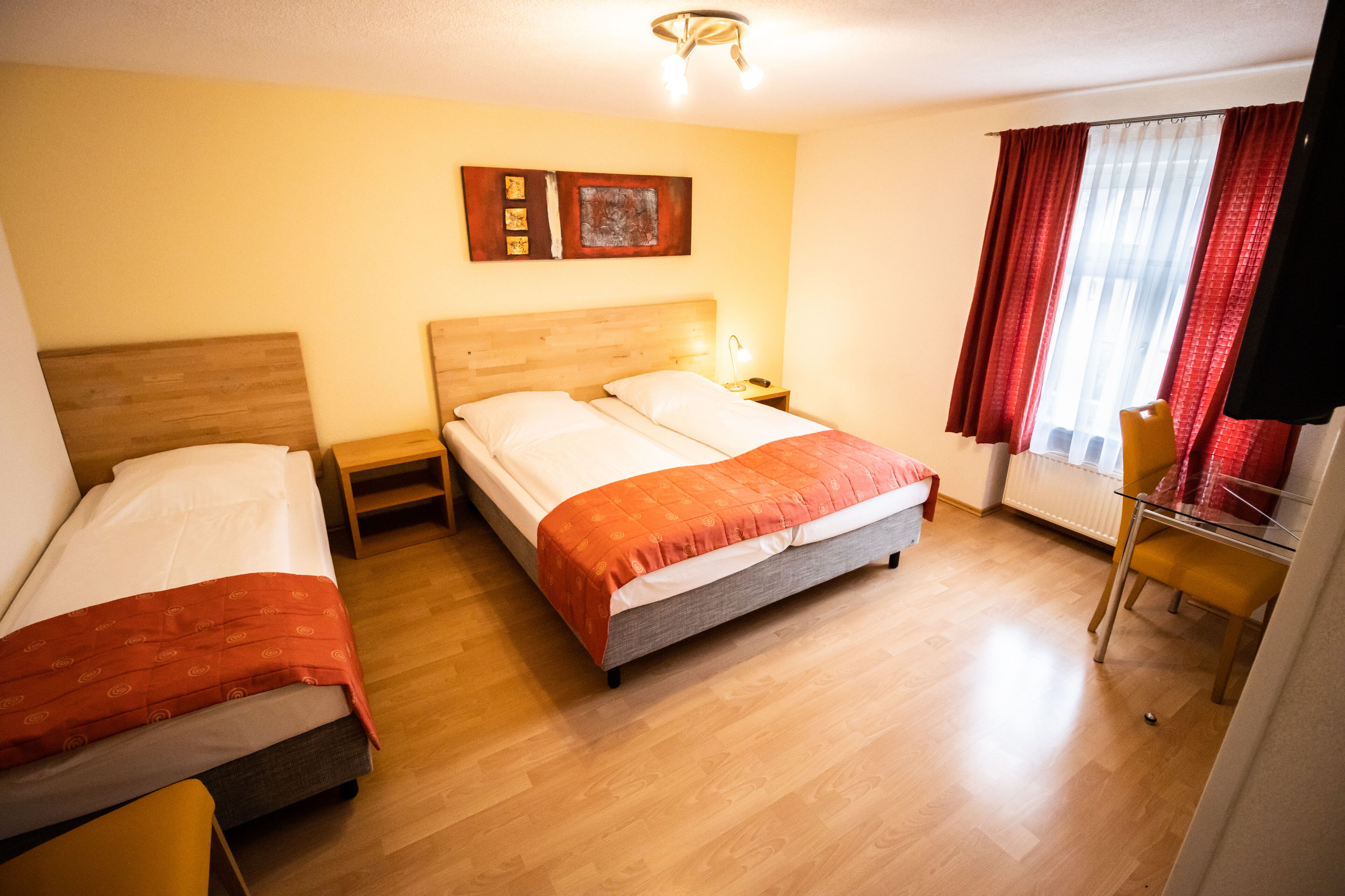 A hotel room with two single beds, a wooden headboard, a nightstand with a lamp, a chair, and a window with red curtains.