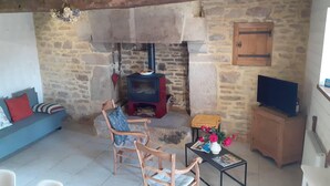 A rustic room with a stone wall, a wooden ceiling, a stone fireplace, a sofa, two wooden chairs, a small table with a vase, and a television on a wooden cabinet. - 17th century house in the heart of a charming little village. (Saint-Servant)