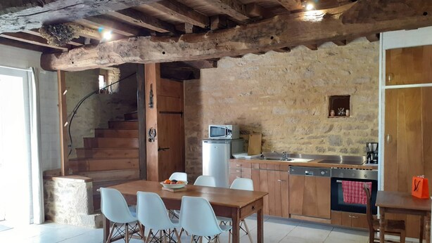 A rustic kitchen with exposed wooden beams, a stone wall, a wooden dining table with chairs, a microwave, a sink, and a stove. - 17th century house in the heart of a charming little village. (Saint-Servant)