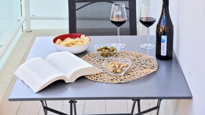 A balcony with a table set for two, including a bottle of wine, glasses, a bowl of snacks, and an open book.