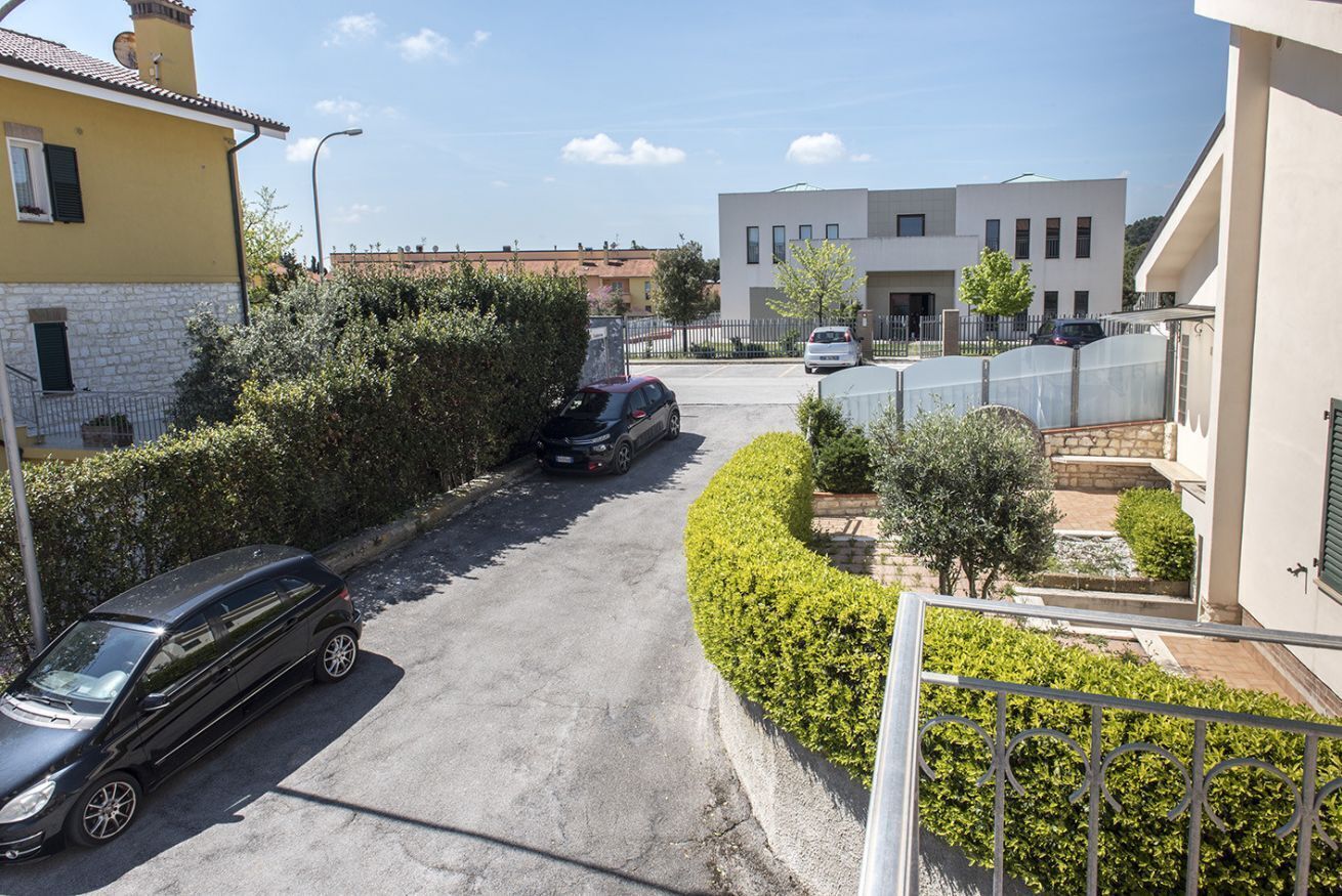 Une rue résidentielle bordée de voitures garées, un immeuble moderne et une haie bien entretenue.