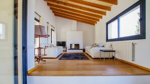 A spacious living room with a fireplace, wooden beams, a large window, and a glass door leading to another room. - Villa Son Paparra in the center of Mallorca. (Montuiri)