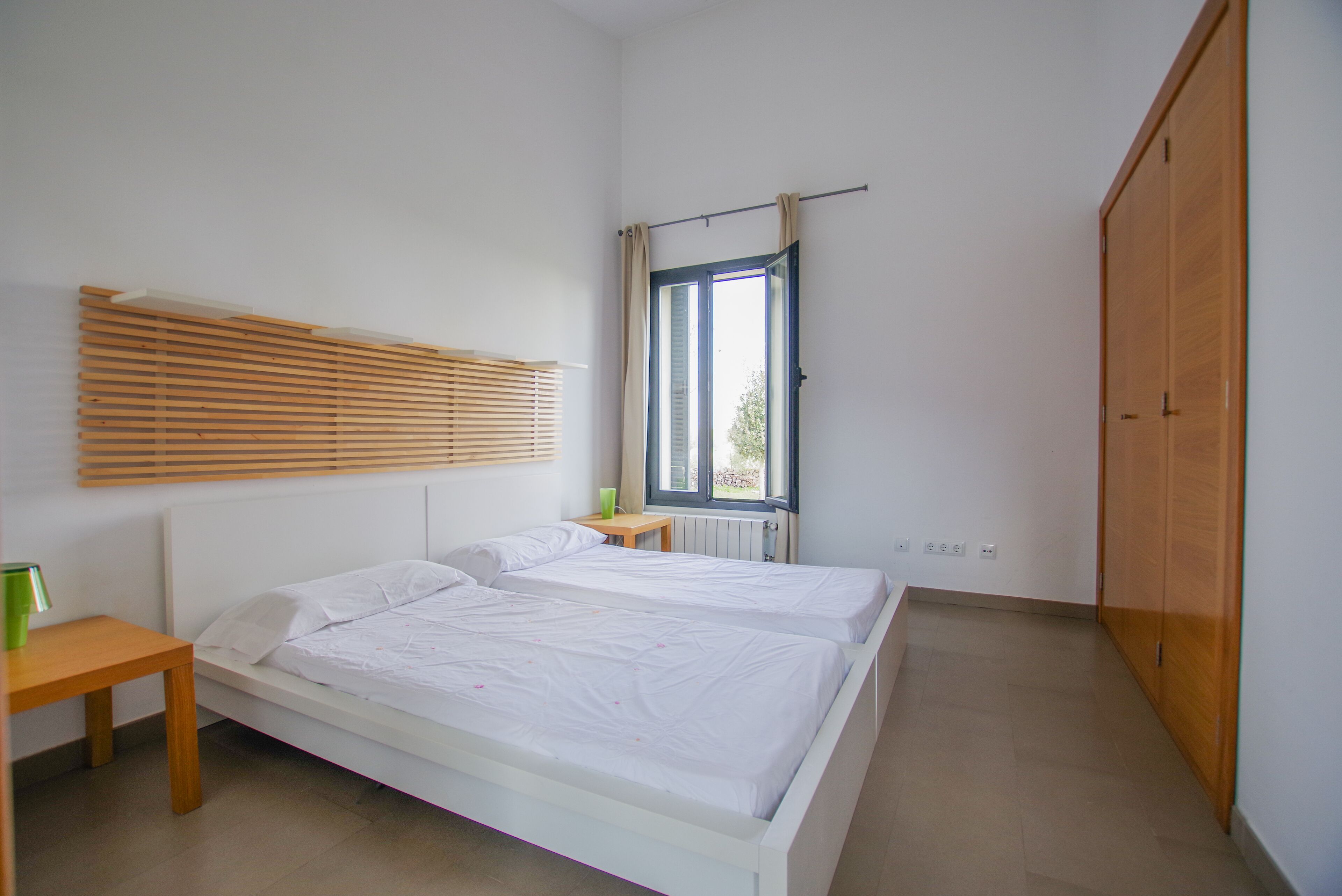 A bedroom with a bed, a window with curtains, a wooden wardrobe, and a bedside table with a lamp.