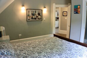 Ein Schlafzimmer mit einem gemusterten Bettbezug, einem Wandbild mit einer Weinflasche und einer Tür zu einer Küche.