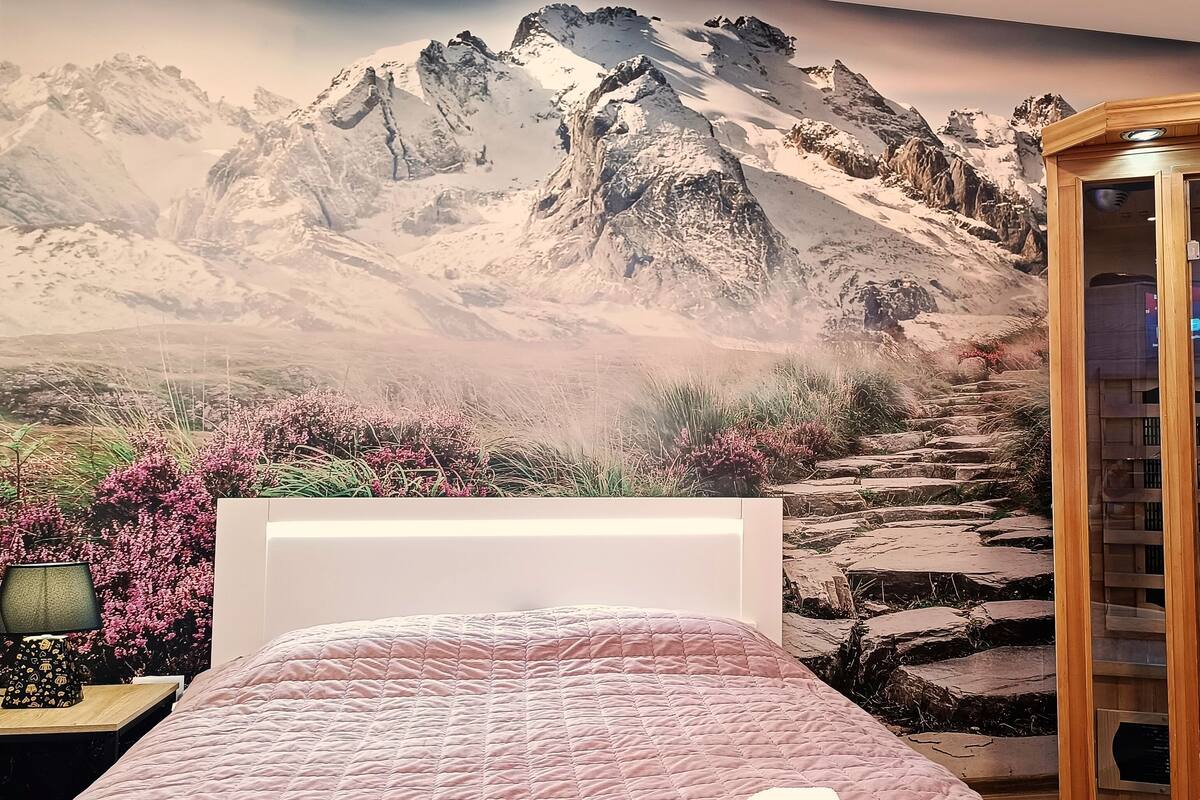 Une chambre avec une fresque représentant une montagne, un lit, une table de chevet avec une lampe et une armoire en bois dotée d’un écran.