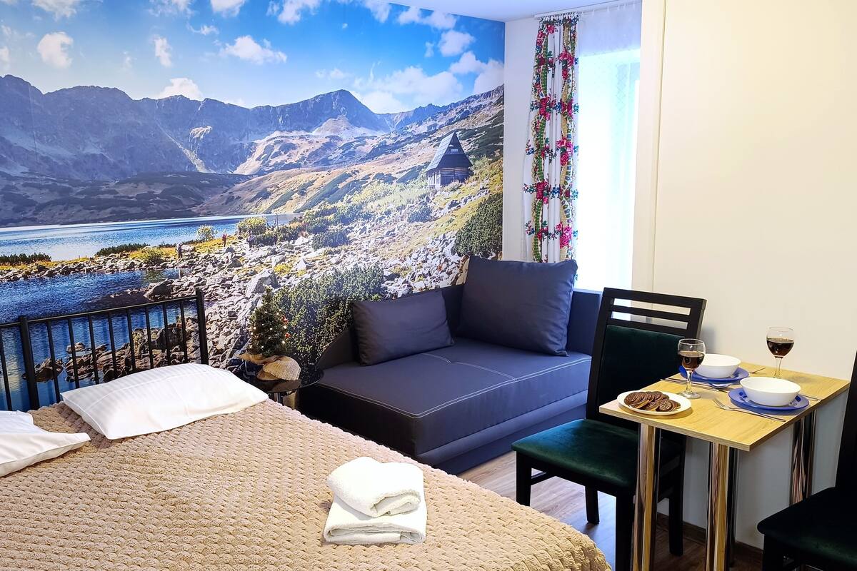 Une chambre avec un lit, un canapé, une table à manger avec une bouteille de vin, et une fresque murale représentant un paysage de montagnes et d’un lac.