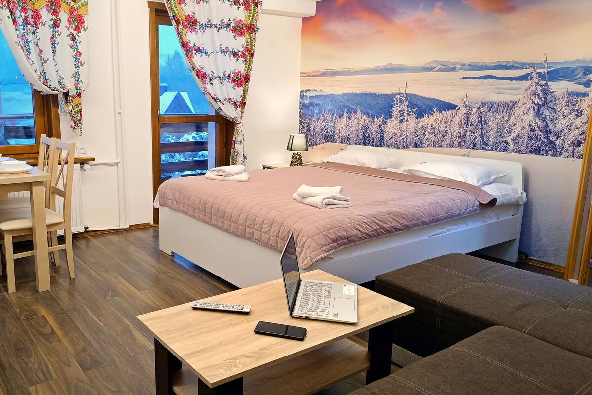 Une chambre d’hôtel avec un lit, un bureau avec un ordinateur portable, une chaise, une table avec une télécommande, et une vue sur des montagnes enneigées par la fenêtre.