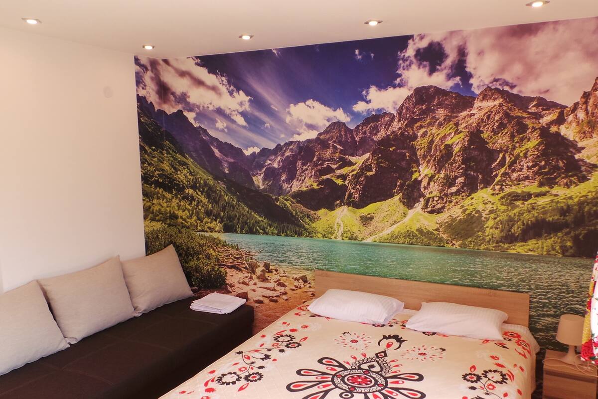 Une chambre avec une fresque murale représentant un paysage de montagne, un lit avec une literie à motifs floraux et un canapé.