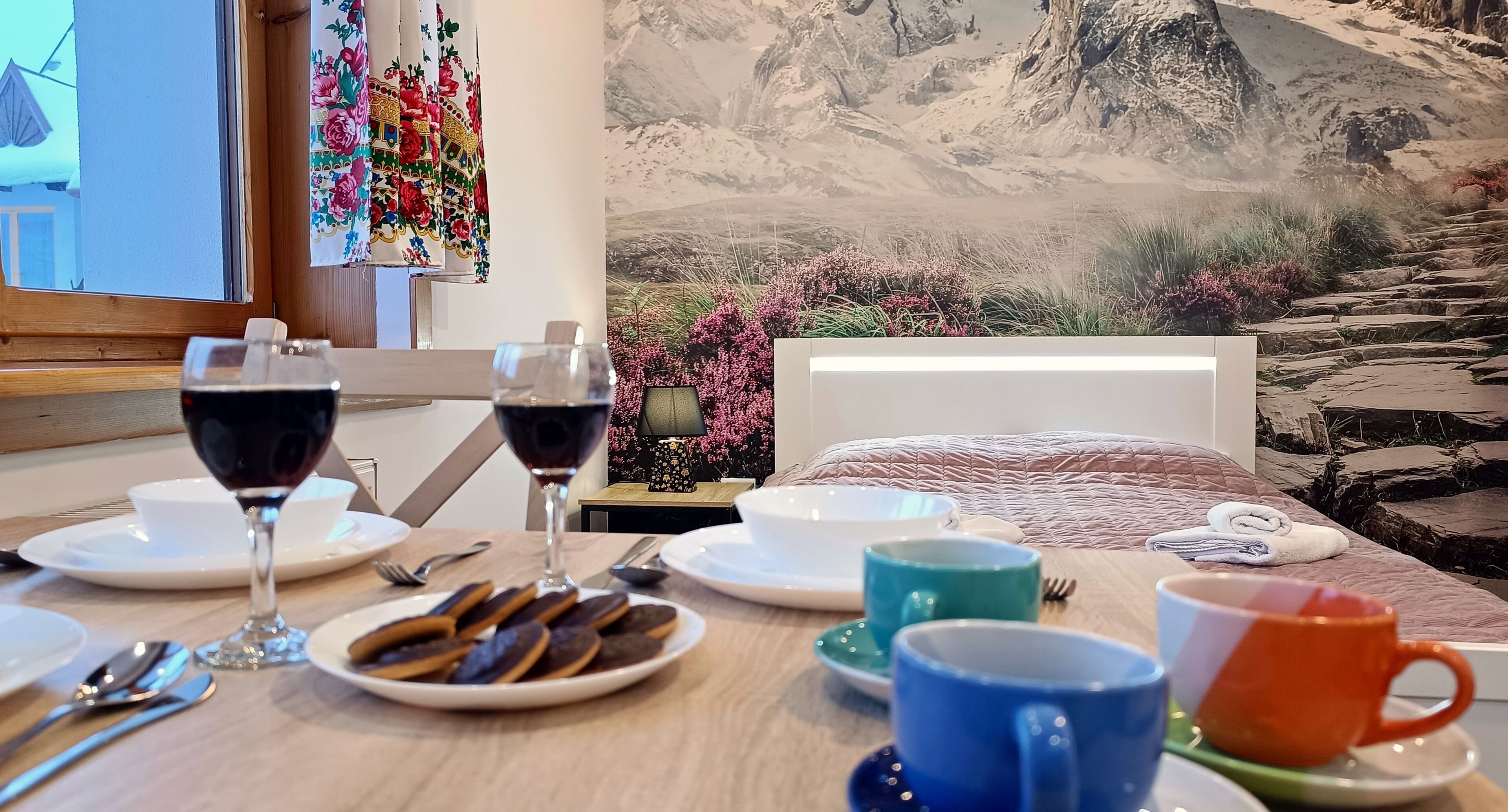 A breakfast table with plates, glasses, and mugs, set against a backdrop of a mountainous landscape.