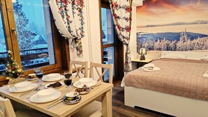 Een knus ingerichte kamer met een bed, een eettafel gedekt voor twee personen en een prachtig uitzicht op besneeuwde bergen door het raam.