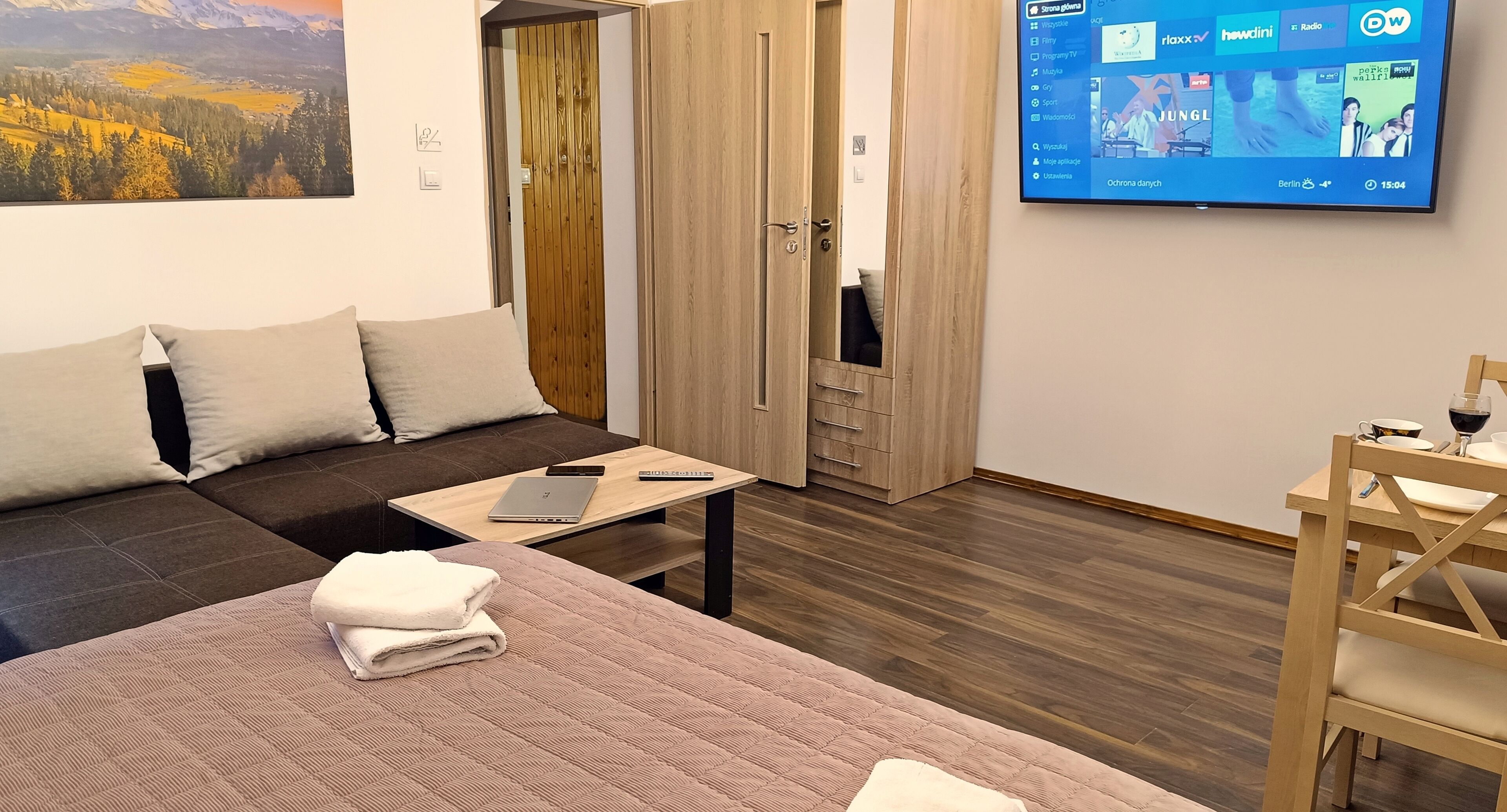 A modern hotel room with a bed, a wooden wardrobe, a television, and a wooden dining table.
