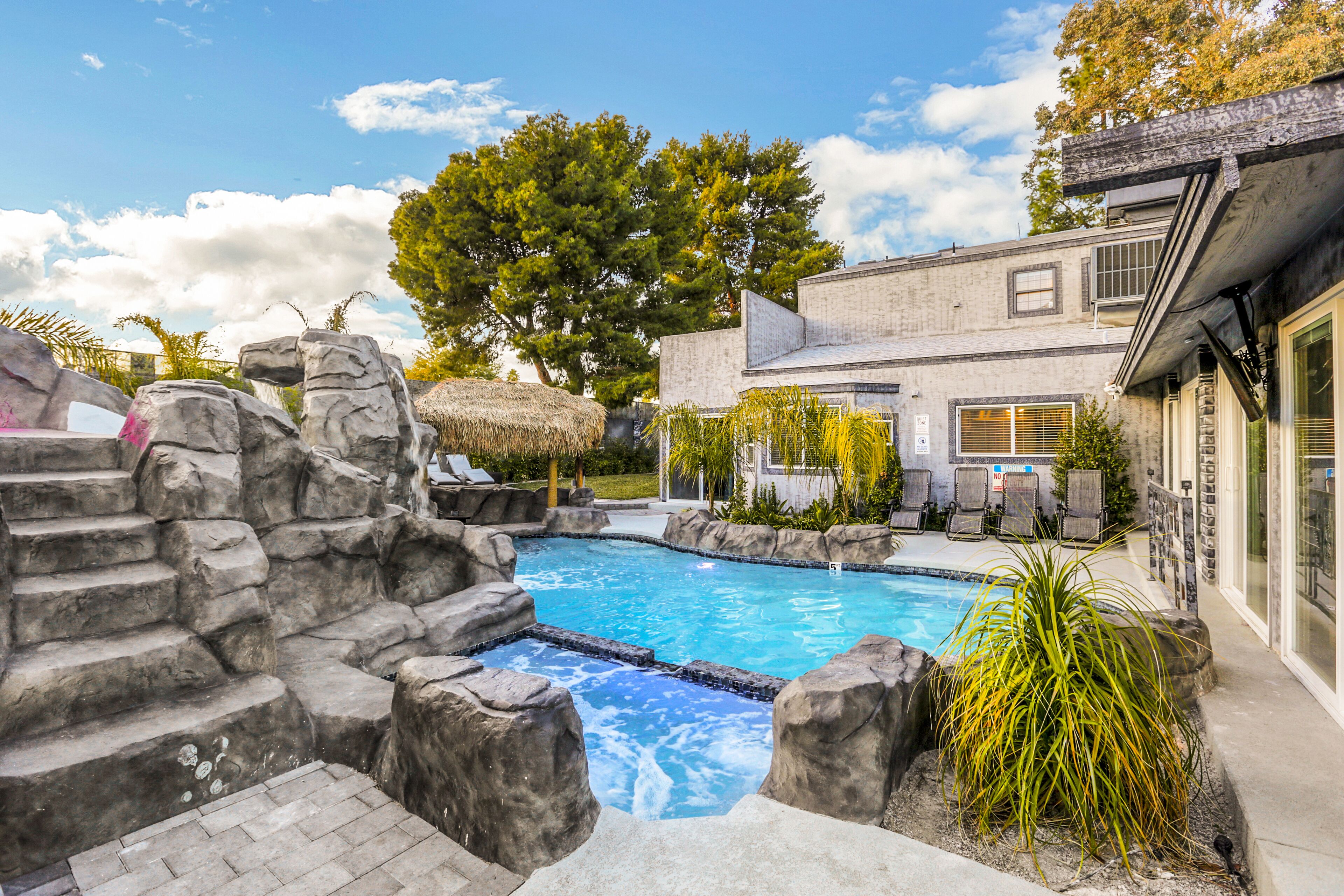 A swimming pool with stone steps and a thatched roof, surrounded by a house with a large tree and outdoor seating.