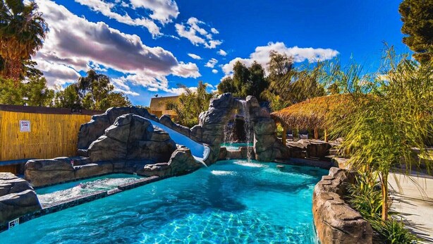 A clear blue pool with a waterfall feature, surrounded by rocks and tropical vegetation.