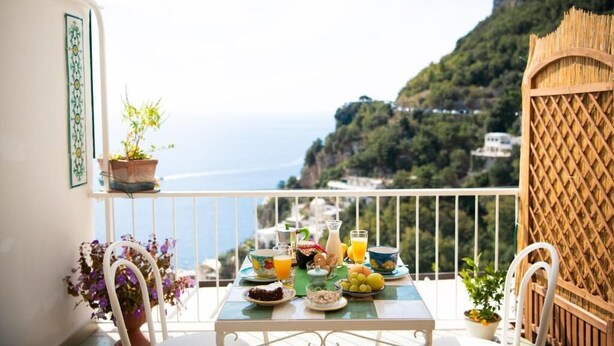 A balcony with a table set for breakfast, overlooking a mountainous coastline.