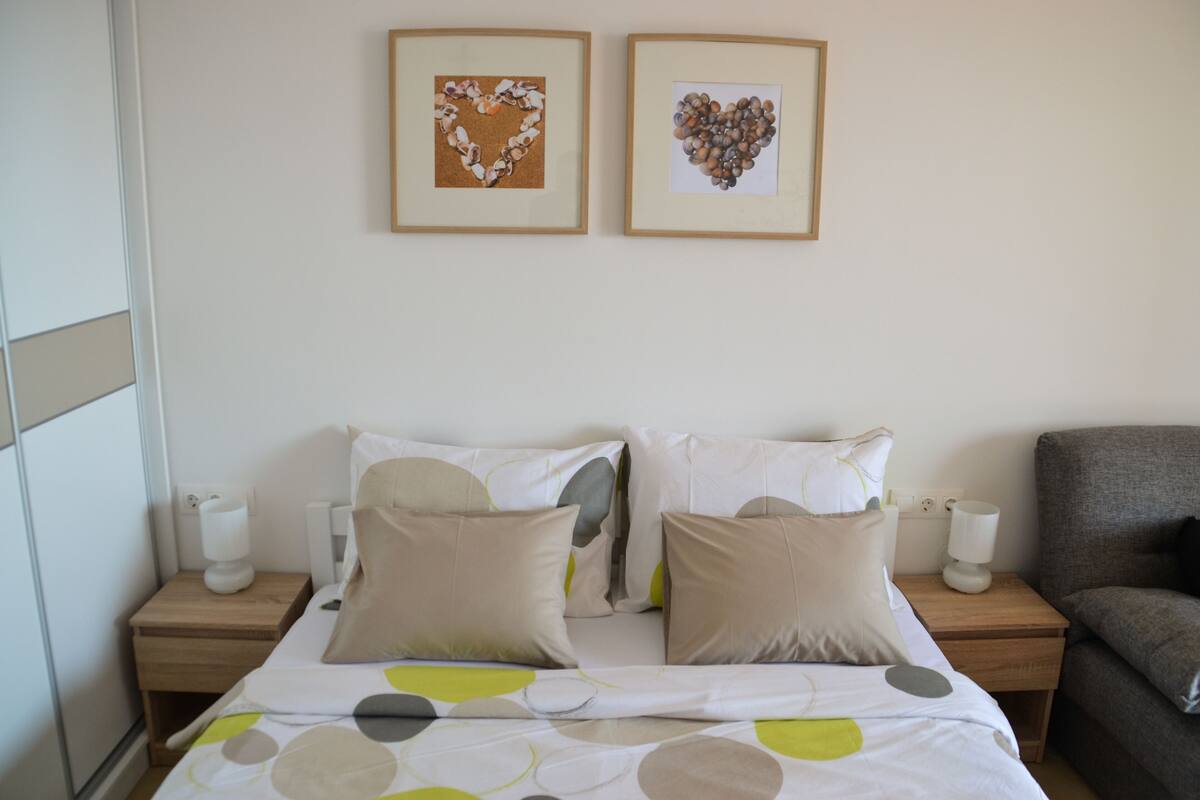 Une chambre à coucher bien rangée, avec un lit, deux tables de chevet, une lampe de chevet et un fauteuil gris.
