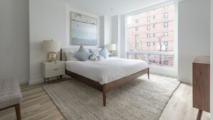 Un dormitorio con una cama grande, una mesita de noche, una silla, una ventana con vistas a edificios y un cuadro en la pared.