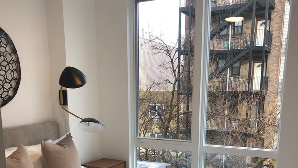 Un dormitorio moderno con una cama grande, una mesita de noche, una lámpara y vistas a un edificio a través de un ventanal amplio.