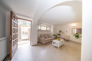 A spacious living room with a beige sofa, a coffee table with fruit, and a view of a hillside through a glass door.