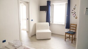 A room with a white chair, a wooden chair, a bed with white linens, a window with blue curtains, and a wall-mounted TV.