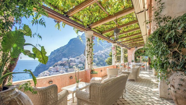 A terrace with wicker furniture, potted plants, and a view of a coastal town.