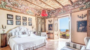 A spacious bedroom with a large bed, a view of the sea, and ornate ceiling artwork.