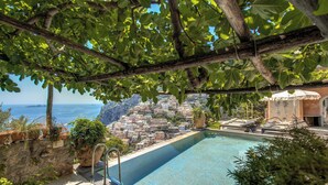 A pool with a view of a coastal town, surrounded by greenery and a pergola.